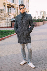A handsome young man in a stylish grey coat, grey jeans and sunglasses stands on the street - 342417450