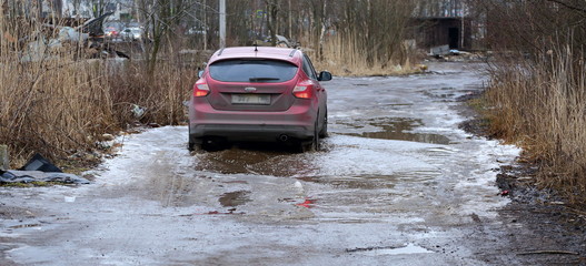 A red car runs over a puddle on a muddy rural road, Dalnevostochniy prospekt, Saint Petersburg, Russia, February 2020