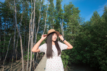 Beautiful young Asian girl smiling to camera enjoy vacation at tropical sea. Teenager girl wearing sun straw hat and white dress standing on wooden bridge with mangrove forest green plant background