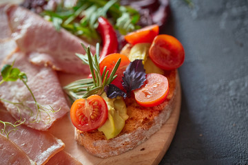 A variety of meat products, laid out on a wooden Board close-up, horizontally, are next to bruschetta with vegetables and fresh herbs
