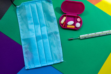Disposable medical masks, a mercury body thermometer and pills lie on a colored surface. The thermometer scale shows the temperature. Selective focus.