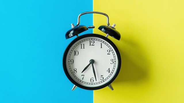 Time lapse Black alarm clock from 7 a.m. to 8 a.m. with bright yellow and blue background and shadows from the changing light of the sun's movements in morning and  working hours concept front shot.