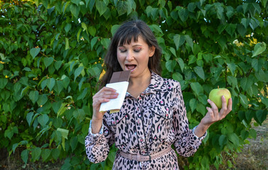 Girl choosing and eating chocolate while holding green fresh apple in other hand. Woman nibbling from sweet and not from fruit 