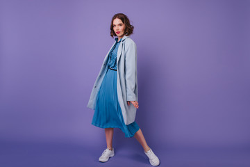 Full-length portrait of refined girl in sneakers and coat. Ecstatic young woman in blue outfit posing with pleasure on purple background.