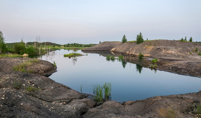 Fototapeta na wymiar Lake in a coal pit with clear water at sunset. Reflection in the water of a coal mountain, hill, trees, reeds.The fuel industry produces coal in an open way.