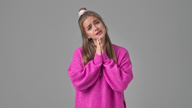 A cute positive young woman is asking for something while saying PLEASE and holding hands like in a praying gesture isolated over a grey background in studio