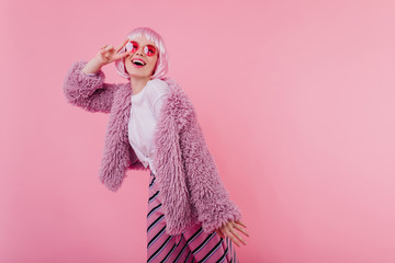 Interested funny girl in periwig and sunglasses posing with peace sign. Indoor shot of blissful...