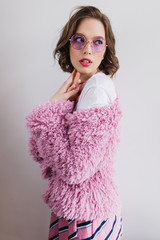 Shapely young woman with brown wavy hair posing in fur jacket. Indoor photo of interested pretty girl in sunglasses and fluffy coat.