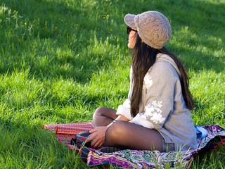 Beautiful Asian woman wearing a hat and sunglasses having a relaxing time in the city park sitting on a colorful blanket surrounded with a green grass.
