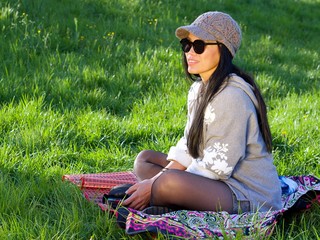 Beautiful Asian woman wearing a hat and sunglasses having a relaxing time in the city park sitting on a colorful blanket surrounded with a green grass. 