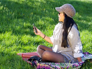 Beautiful Asian woman wearing a hat and sunglasses having a relaxing time in the city park sitting on a colorful blanket surrounded with a green grass using her phone making a selfie.
