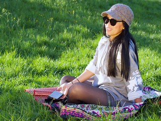Beautiful Asian woman wearing a hat and sunglasses having a relaxing time in the city park sitting on a colorful blanket surrounded with a green grass. 
