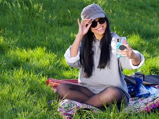 Beautiful Asian woman wearing a hat and sunglasses having a relaxing time in the city park sitting on a colorful blanket surrounded with a green grass using her phone making a selfie.