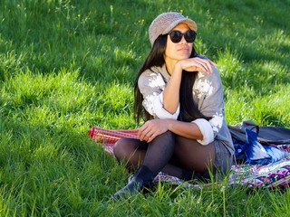 Beautiful Asian woman wearing a hat and sunglasses having a relaxing time in the city park sitting on a colorful blanket surrounded with a green grass.