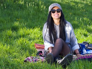 Beautiful Asian woman wearing a hat and sunglasses having a relaxing time in the city park sitting on a colorful blanket surrounded with a green grass. 