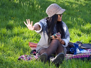 Beautiful Asian woman wearing a hat and sunglasses having a relaxing time in the city park sitting on a colorful blanket surrounded with a green grass. 