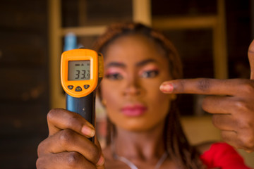 Close-up shot of a young african woman holding infrared forehead thermometer (thermometer gun) to...