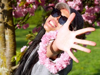 Portrait of a beautiful Asian woman wearing a hat and sunglasses holding a cherry blossoms flower circle in the city park next to a cherry tree enjoying the sun.