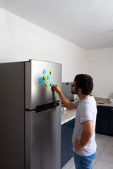 Hispanic young man taking and reading sticky posticks in his fridge