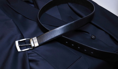 A black leather belt with steel fittings lies horizontally on a black men's jacket. An expensive...