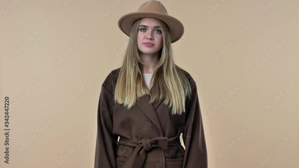 Poster A young pretty woman wearing hat and coat is showing a thumb down gesture isolated over the beige background in studio