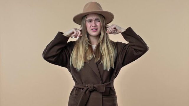A young displeased woman wearing hat and coat is covering her ears and showing nonsense content isolated over the beige background in studio