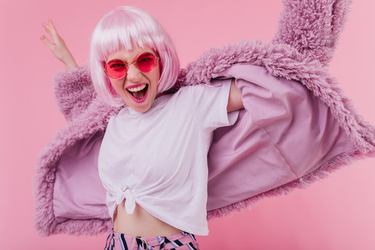Excited stylish girl in sunglasses and wig posing on pink background. Studio photo of blissful young woman dancing and laughing.