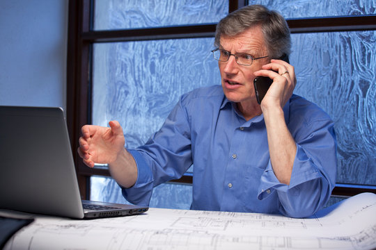 Mature angry businessman talking on mobile phone and looking at the monitor of a laptop