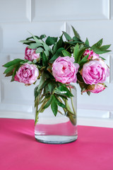 Bouquet of pink peonies in glass vase against white background and wooden table. Mothers day card. Beautiful peony flower for catalog or online store. Floral shop and delivery concept. Copy space.