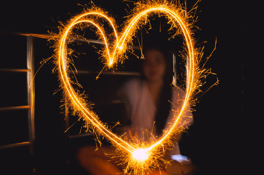 Close-up Of Heart Shaped Light Painting Against Woman At Night