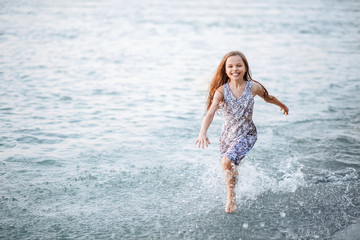 A happy young girl runs along the shore on the water and waves her hat. The girl is happy with the onset of summer