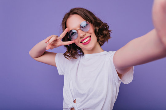 Pleasant girl with tattoo making selfie in studio and laughing. Good-looking young woman with brown wavy hair taking picture of herself on bright purple background.