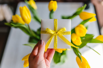 Woman hand hold gift box on yellow tulips background. Space for message. Greeting card. Holiday greeting card for Valentine's, Women's, Mother's Day, Easter! Happy Birthday. Top view, flat lay.