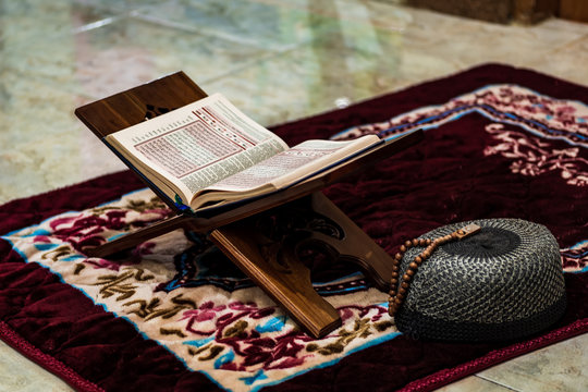 Quran, the Scripture of Muslims on rihal or wood book stand, prayer beads or tasbeeh, skullcap hat, at home.  The concept of staying home in ramadan during a corona virus disease pandemic situation.
