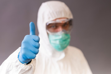 Male doctor with surgical mask, goggles and protective suit pointing okay on gray backround.