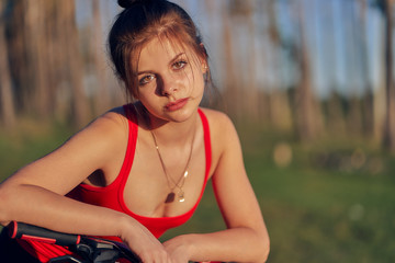 Cute girl with pendant on a chain in a red top with her hair tied in a bun, leaned on the handlebars of a bicycle. sunset on nature against the background of the forest and trees. Close-up.