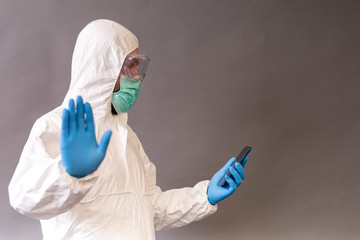 Doctor with surgical mask, goggles and protective suit holding his phone and showing stop  with a hand up on gray backround.