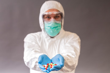 Male doctor with surgical mask, goggles and protective suit holding stack of many different pills.