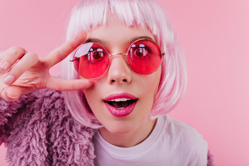 Close-up shot of funny european girl in colorful periwig and pink sunglasses. Indoor photo of...
