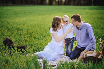 Fototapeta premium Family on a picnic in the forest. In the meadow. Picnic basket with food. The dog runs and wants to play. A child with glasses. Blue color in clothes. To eat outdoors. Time with family.