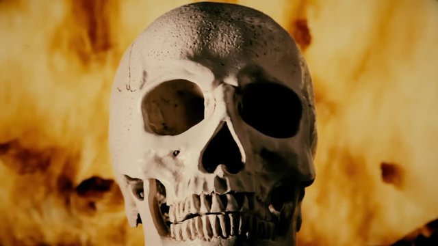 A front close-up shot of a scary human skull, standing still, eyes facing the viewer, over wild flames from a big fire pyre.
