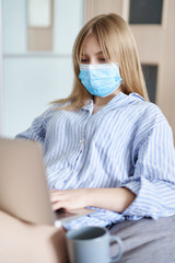 Concentrated freelance with distance work during coronavirus pandemic. Vertical photo of young woman sitting on a beanbag in medical mask with laptop in front of her studying or working.