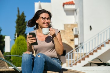 Mature woman relaxing in resort, sitting in chair with cup of coffee and smartphone