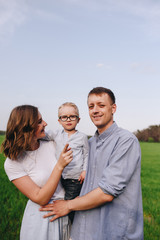 Fototapeta premium Family in the forest for a walk. Picnic in the meadow, on the green grass. Blue clothes. Mom, dad, son. Son on the hands of dad. A pet. Black Dog. The boy with glasses.