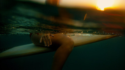 HALF UNDERWATER: Surfer girl sits on surfboard and waits for a wave at sunset.
