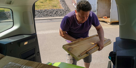 Plump,dark-haired,middle-aged man unloads wooden planks from trunk of compact MPV to dump it at station of collection of large-size garbage in Germany,Europe.Spring sunny day.Selective focus,close up