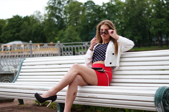 Sexy woman in a red skirt posing outdoors in the park.A bitchy girl smokes a cigarette and catches her eye.Beautiful model in various poses. Summer nature,all in green and red colors.Glasses on a girl