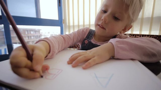 Child draws with colored pencils. Blonde caucasian girl draws character B with colored pencil on black table at home. Close-up.