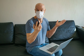 Man is sitting on a sofa in a protective mask with a laptop and phone, remote work in quarantine