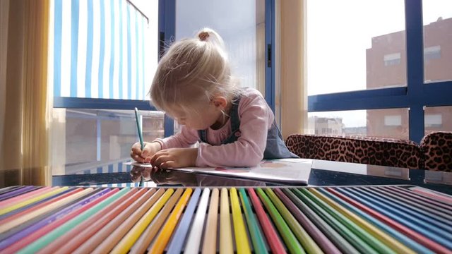 Child draws with colored pencils. Blonde caucasian girl with pigtail draws with pencil on background. Tin box with color pencils on foreground.